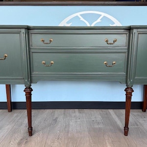 May include: A green painted sideboard with two drawers and two cabinet doors. The drawers and doors have brass-colored handles. The sideboard has four decorative, dark brown legs.