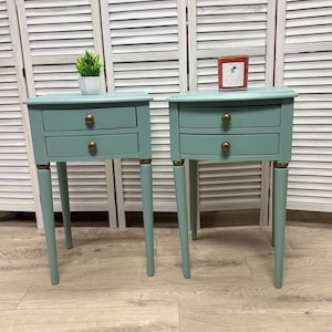 May include: Two matching sage green bedside tables, each featuring two drawers with round brass-toned knobs. The tables have four tapered legs with brass-toned accents at the top. A small green plant in a white pot sits on one, and a red candle on the other.