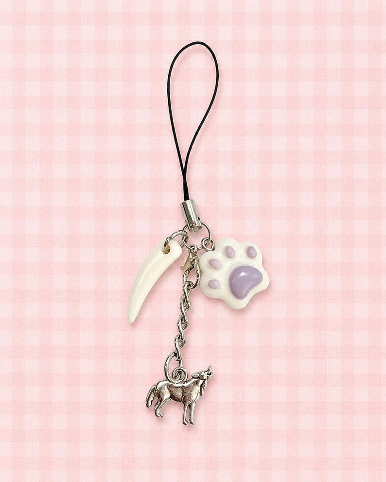 May include: A phone charm featuring a silver-toned metal chain with three dangling pendants: a white curved tusk-shaped charm, a white and light purple paw print charm, and a silver-toned metal wolf charm, all attached to a black cord loop.