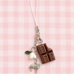 May include: A phone charm featuring a brown chocolate bar pendant with the word MILK embossed on it, a small white bell-shaped flower with a green leaf, and a silver-toned wolf charm, all attached to a white cord with a silver metal connector.