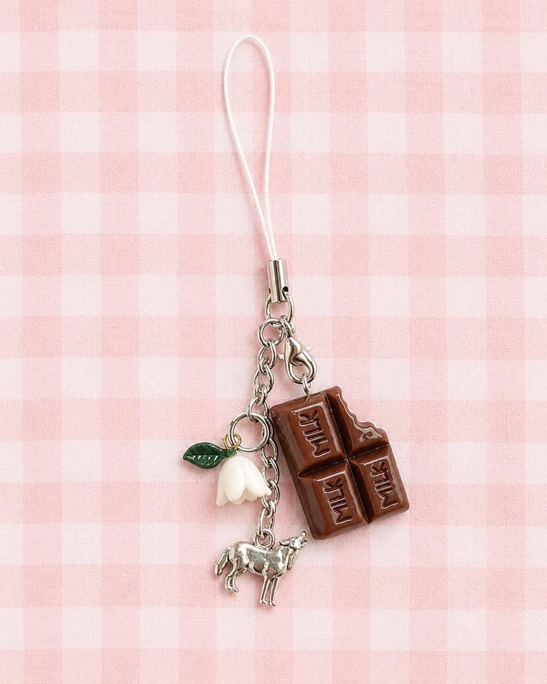 May include: A phone charm featuring a brown chocolate bar pendant with the word MILK embossed on it, a small white bell-shaped flower with a green leaf, and a silver-toned wolf charm, all attached to a white cord with a silver metal connector.