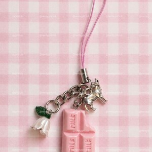 May include: A phone charm featuring a pink strap, a silver-toned metal chain, a silver-toned wolf charm, a white flower charm with a green leaf, and a pink chocolate bar charm with the word MILK embossed on the surface of the chocolate segments.