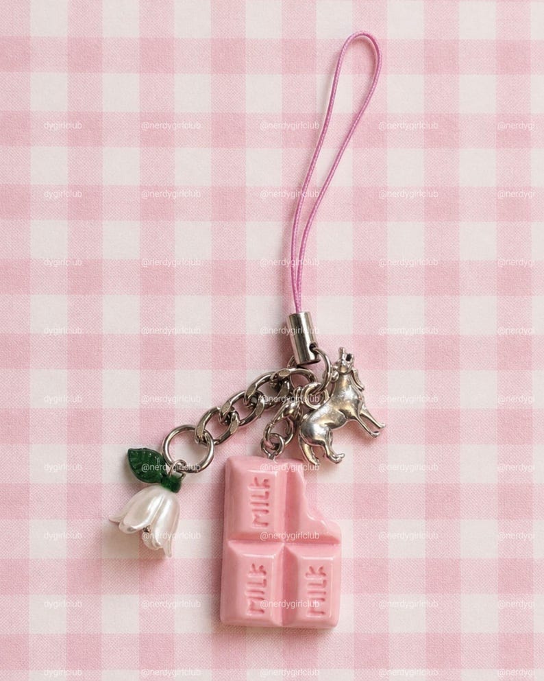 May include: A phone charm featuring a pink strap, a silver-toned metal chain, a silver-toned wolf charm, a white flower charm with a green leaf, and a pink chocolate bar charm with the word MILK embossed on the surface of the chocolate segments.
