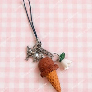 May include: A phone charm featuring a dark blue cord attached to a silver-toned metal chain. The charm includes a silver-colored wolf figurine, a brown textured ice cream cone, and a small white flower with a green leaf attached to the chain.