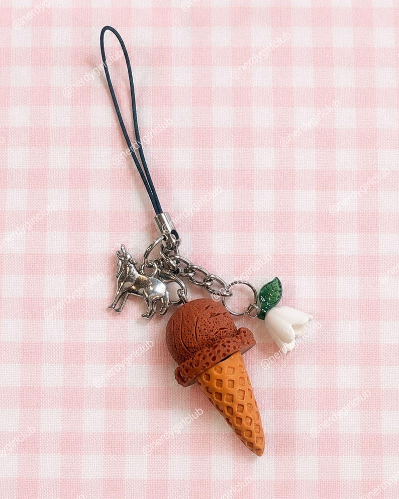 May include: A phone charm featuring a dark blue cord attached to a silver-toned metal chain. The charm includes a silver-colored wolf figurine, a brown textured ice cream cone, and a small white flower with a green leaf attached to the chain.