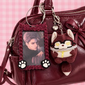May include: A dark red glossy handbag featuring a rectangular photo frame keychain with a ruffled border and two white paw prints, alongside a plush fox-like character keychain wearing black glasses and a yellow neck ribbon attached to a silver chain.