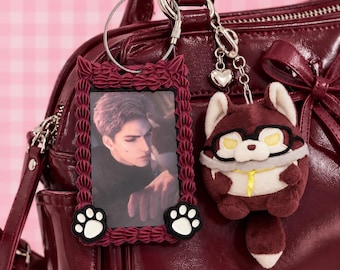 May include: A dark red glossy handbag featuring a rectangular photo frame keychain with a ruffled border and two white paw prints, alongside a plush fox-like character keychain wearing black glasses and a yellow neck ribbon attached to a silver chain.