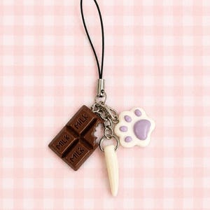 May include: A phone charm featuring three dangling pendants attached to a black cord: a brown chocolate bar charm with the word MILK embossed on it, a white fang-shaped charm, and a white paw print charm with a light purple center pad.