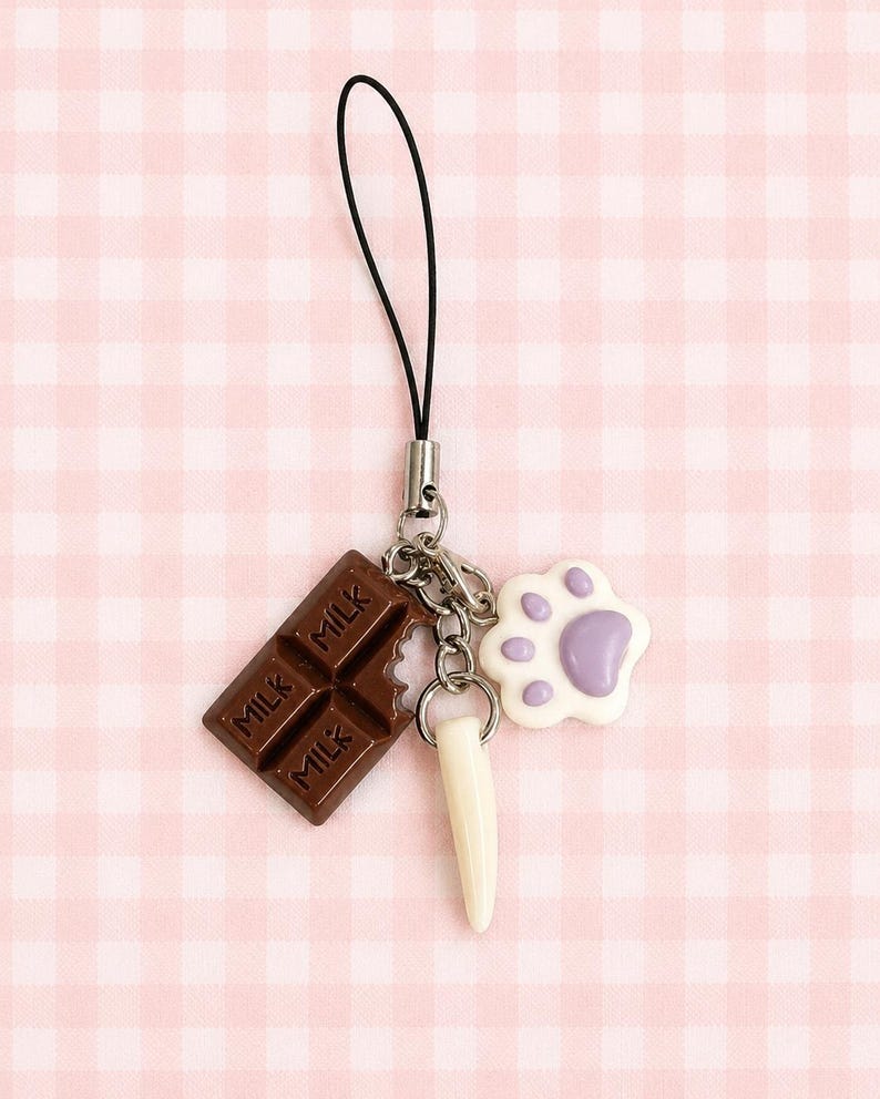 May include: A phone charm featuring three dangling pendants attached to a black cord: a brown chocolate bar charm with the word MILK embossed on it, a white fang-shaped charm, and a white paw print charm with a light purple center pad.