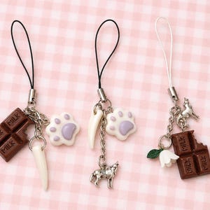 May include: Three phone charms featuring silver-tone hardware and black or white cords. The charms include miniature brown chocolate bars with the word MILK, white and purple paw prints, white curved spikes, a silver wolf, and a white flower.