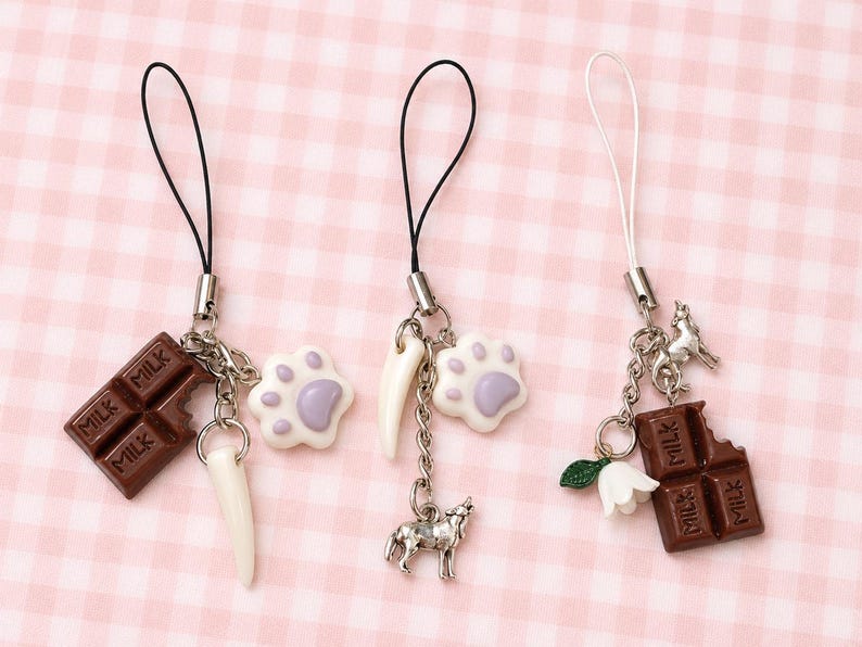 May include: Three phone charms featuring silver-tone hardware and black or white cords. The charms include miniature brown chocolate bars with the word MILK, white and purple paw prints, white curved spikes, a silver wolf, and a white flower.