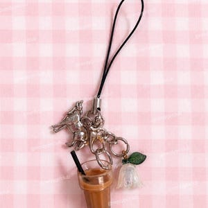 May include: A charm strap featuring a silver-toned wolf figurine, a miniature plastic iced coffee cup with a black straw, and a translucent white flower charm with a green leaf, all attached to a black cord with a silver metal connector.