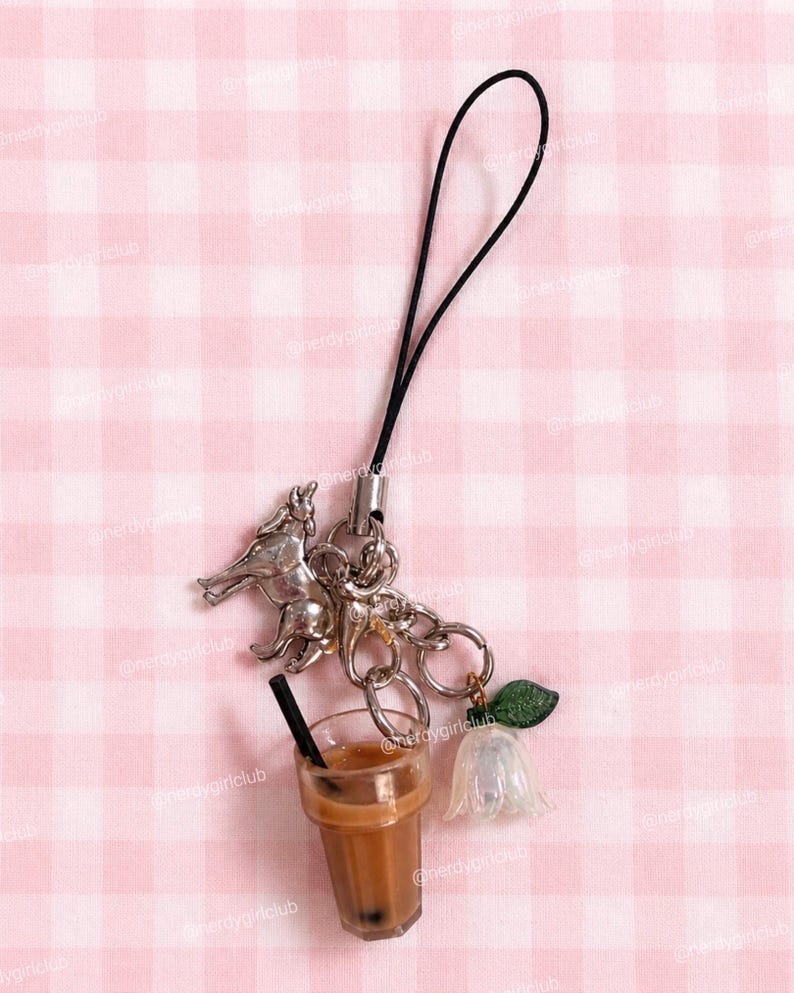 May include: A charm strap featuring a silver-toned wolf figurine, a miniature plastic iced coffee cup with a black straw, and a translucent white flower charm with a green leaf, all attached to a black cord with a silver metal connector.