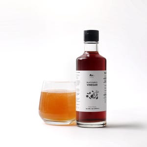 May include: A bottle of black garlic vinegar with a black cap and a white label, next to a glass of amber-colored liquid. The label reads "Black Garlic Vinegar" with illustrations of garlic cloves. The bottle contains 16.9 fl oz (500ml).