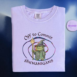 May include: A light purple Comfort Colors t-shirt with the text "Off to Commit Shenanigans" and a cartoon frog wizard graphic. The shirt has a crew neck and short sleeves. A small round color swatch labeled "Orchid" is in the upper right corner.