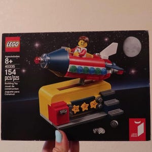 Puede incluir: Un juego de construcción LEGO con un cohete rojo, azul y blanco con una minifigura, sobre un fondo estrellado con una luna. La caja dice "LEGO" e incluye el número de juego 40335 y 154 piezas.