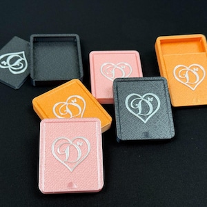 Peut inclure: Plusieurs petites boîtes carrées de différentes couleurs, dont le noir, l'orange et le rose. Chaque boîte est ornée d'un motif en forme de cœur avec la lettre "D" et un papillon. Les boîtes semblent être faites d'un matériau texturé.