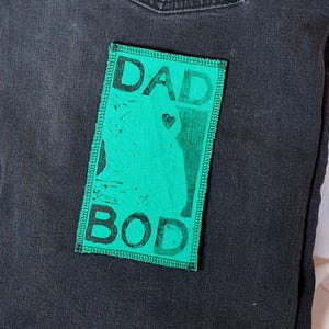 Könnte beinhalten: Schwarze Jeans mit einem grünen Aufnäher mit den weißen Worten "DAD BOD". Der Aufnäher zeigt eine Silhouette einer Person. Die Jeans hat eine Gesäßtasche.