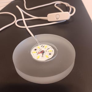 May include: A frosted gray LED light with a white cord and a small on/off switch. The light has a circular design with a central array of small, bright LED bulbs. The cord is white and appears to be made of a flexible material.