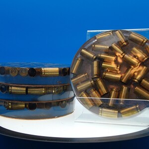 May include: Two resin art pieces. One is a stack of three cylinders with brass bullet casings embedded in the sides. The other is a clear, round resin piece with numerous brass bullet casings suspended inside. Both are displayed on a white surface.