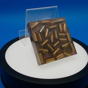 May include: A rectangular block with a brown surface embedded with numerous brass-colored bullet casings. One silver-colored casing is also visible. The block is displayed on a white and black rotating stand against a blue backdrop.