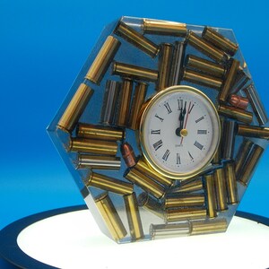 May include: A unique hexagonal clock encased in clear resin, featuring a collection of brass and copper bullet casings. The clock face has Roman numerals and gold-colored hands. The clock is set against a bright blue background.