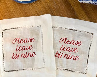 Puede incluir: Dos posavasos de lino blanco con texto bordado en rojo que dice "Please leave by nine". Cada posavasos tiene un borde cosido. Piezas decorativas de cerámica azul y blanca en el fondo.