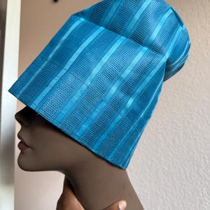 May include: A bright turquoise blue hat with a structured, tall, cylindrical shape. The fabric features a repeating pattern of vertical stripes alternating between smooth, shiny bands and textured, woven sections that create a subtle depth.