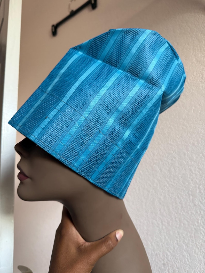 May include: A bright turquoise blue hat with a structured, tall, cylindrical shape. The fabric features a repeating pattern of vertical stripes alternating between smooth, shiny bands and textured, woven sections that create a subtle depth.
