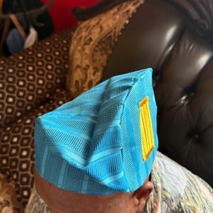 May include: A bright turquoise blue structured cap featuring a textured, striped fabric pattern and a rectangular yellow accent patch on the side. The headwear has a distinct geometric shape with sharp, defined edges and a firm, upright construction.
