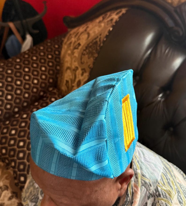 May include: A bright turquoise blue structured cap featuring a textured, striped fabric pattern and a rectangular yellow accent patch on the side. The headwear has a distinct geometric shape with sharp, defined edges and a firm, upright construction.