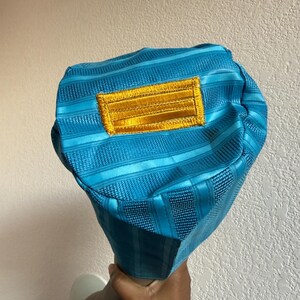 May include: A bright turquoise bucket hat featuring a textured, striped fabric pattern and a rectangular, embroidered gold patch centered on the front. The hat has a structured, rounded crown and a short, downward-sloping brim.