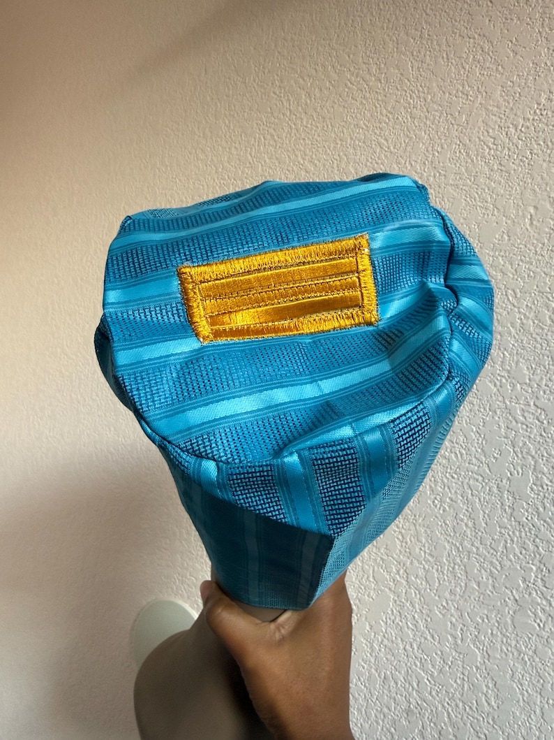 May include: A bright turquoise bucket hat featuring a textured, striped fabric pattern and a rectangular, embroidered gold patch centered on the front. The hat has a structured, rounded crown and a short, downward-sloping brim.