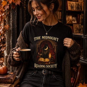 May include: A black t-shirt featuring the text THE MIDNIGHT READING SOCIETY in gold lettering. The graphic depicts an open book, a lit candle, and a crescent moon visible through an arched window with red curtains and a small butterfly.