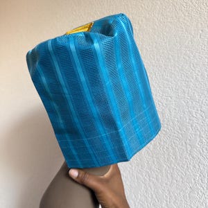 May include: A cylindrical teal blue hat featuring a textured vertical striped pattern and a small, rectangular gold-colored accent patch on the top edge. The hat has a structured, stiff appearance with a flat top and straight sides.
