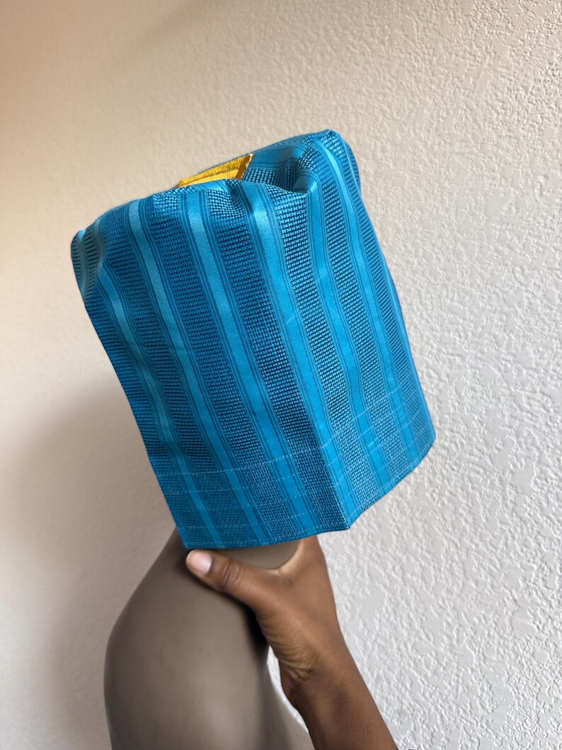 May include: A cylindrical teal blue hat featuring a textured vertical striped pattern and a small, rectangular gold-colored accent patch on the top edge. The hat has a structured, stiff appearance with a flat top and straight sides.