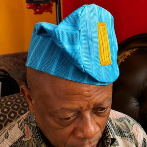 May include: A bright turquoise blue cap featuring a structured, folded design with vertical textured stripes and a prominent, rectangular mustard yellow embroidered accent patch on the side. The fabric has a subtle sheen and a woven appearance.