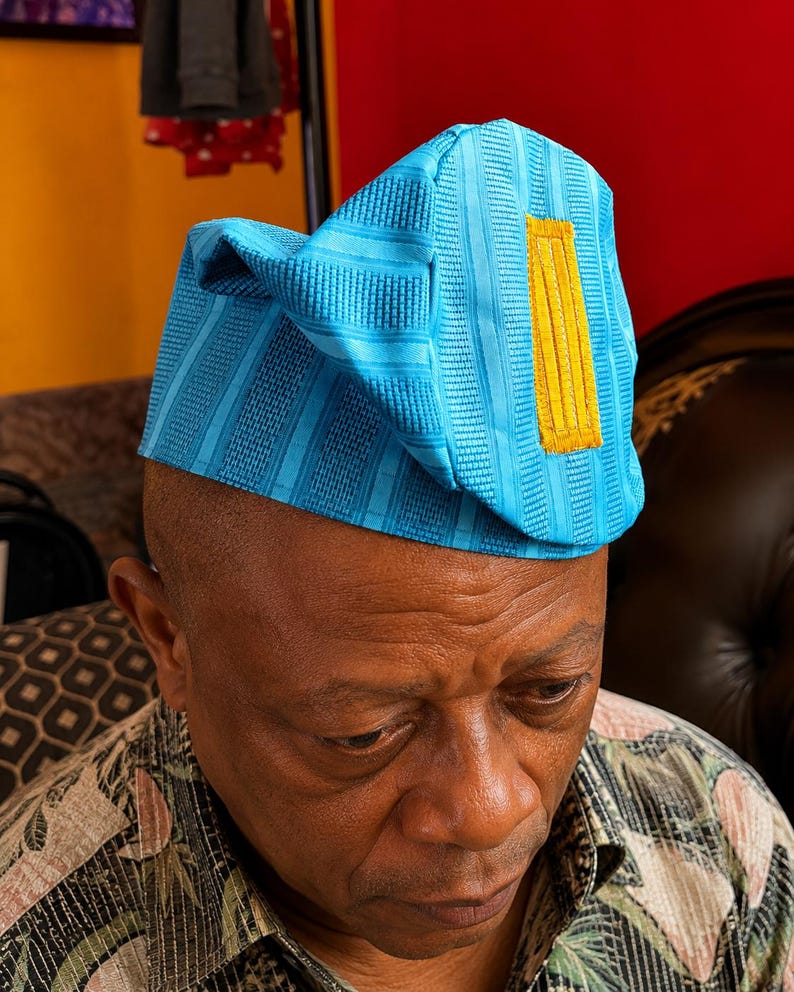 May include: A bright turquoise blue cap featuring a structured, folded design with vertical textured stripes and a prominent, rectangular mustard yellow embroidered accent patch on the side. The fabric has a subtle sheen and a woven appearance.