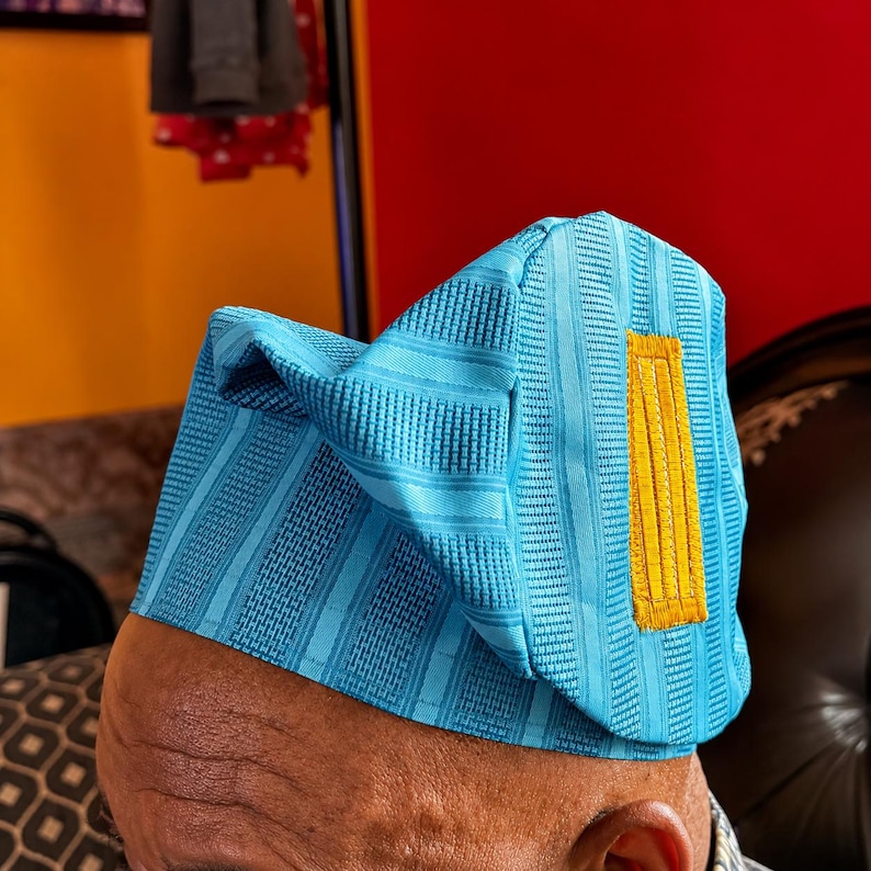 May include: A light blue structured cap featuring a textured, striped fabric pattern and a prominent, rectangular mustard yellow embroidered patch on the side. The headwear has a distinct, angular, folded design with a stiffened, upright silhouette.