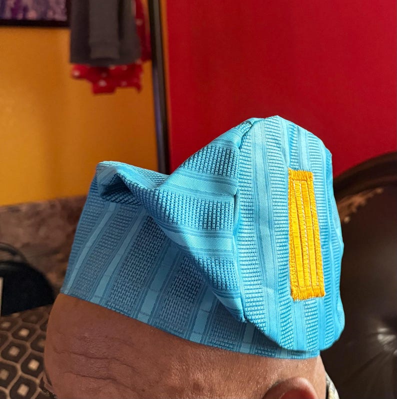 May include: A light blue structured cap featuring a textured, striped fabric pattern and a prominent, rectangular yellow embroidered accent on the side. The hat has a distinct, angular shape with a folded top section and a firm, upright brim.