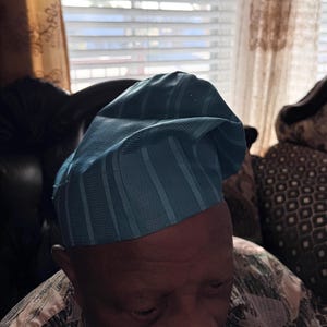 May include: A light blue traditional cap featuring a structured, rounded crown with vertical textured stripes. The fabric has a subtle sheen and a folded, asymmetrical design that sits atop the head, showcasing a consistent woven pattern.
