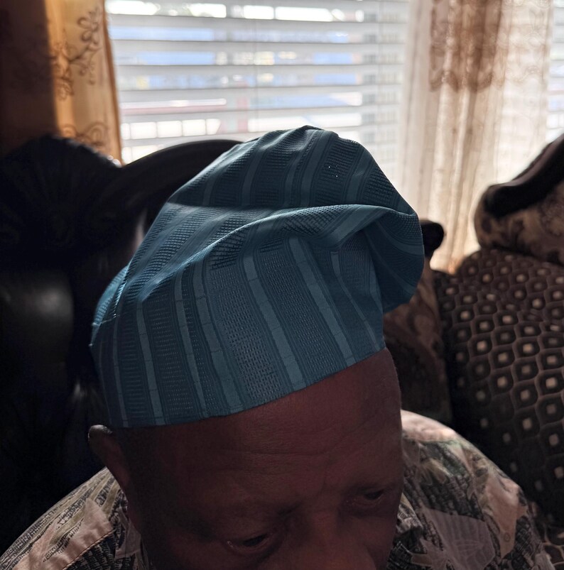May include: A light blue traditional cap featuring a structured, rounded crown with vertical textured stripes. The fabric has a subtle sheen and a folded, asymmetrical design that sits atop the head, showcasing a consistent woven pattern.