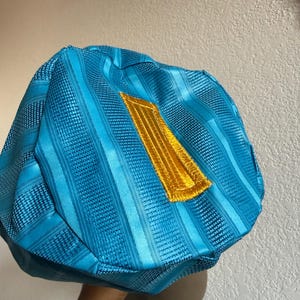 May include: A bright turquoise blue cap featuring a structured, multi-paneled design with alternating smooth and textured vertical stripes. A prominent, rectangular gold-colored embroidered patch is centered on the front of the cap.