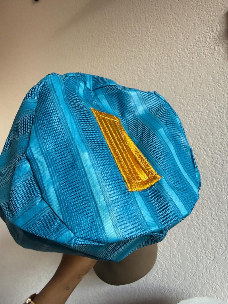 May include: A bright turquoise blue cap featuring a structured, multi-paneled design with alternating smooth and textured vertical stripes. A prominent, rectangular gold-colored embroidered patch is centered on the front of the cap.