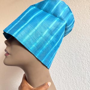 May include: A bright turquoise blue cap featuring a structured, stiffened design with alternating vertical stripes of smooth fabric and textured, woven patterns. The headwear has a tall, slightly angled silhouette that sits firmly on the head.