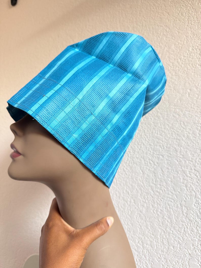 May include: A bright turquoise blue cap featuring a structured, stiffened design with alternating vertical stripes of smooth fabric and textured, woven patterns. The headwear has a tall, slightly angled silhouette that sits firmly on the head.
