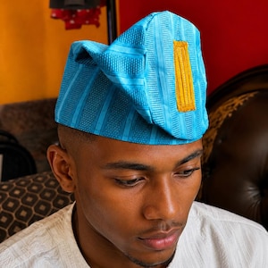 May include: A bright turquoise blue cap featuring a structured, folded design with vertical textured stripes and a prominent, rectangular mustard yellow patch centered on the front panel. The fabric has a subtle sheen and a consistent woven pattern.
