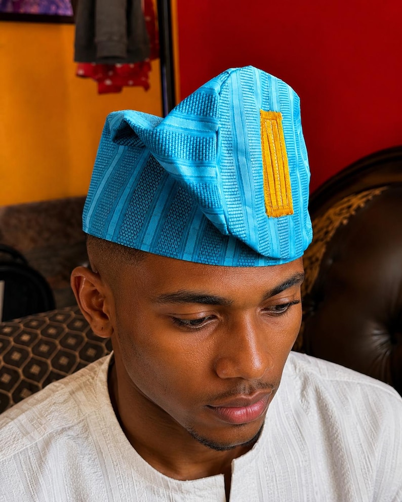 May include: A bright turquoise blue cap featuring a structured, folded design with vertical textured stripes and a prominent, rectangular mustard yellow patch centered on the front panel. The fabric has a subtle sheen and a consistent woven pattern.