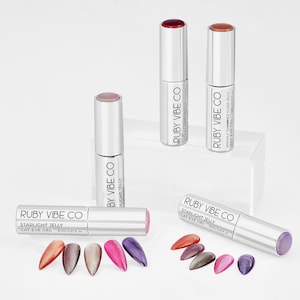 May include: A collection of Ruby Vibe Co. Starlight Jelly Cat Eye Gel nail polish in various colors. The image shows several silver tubes with colored caps and swatches of the nail polish in shades of red, pink, purple, and brown.