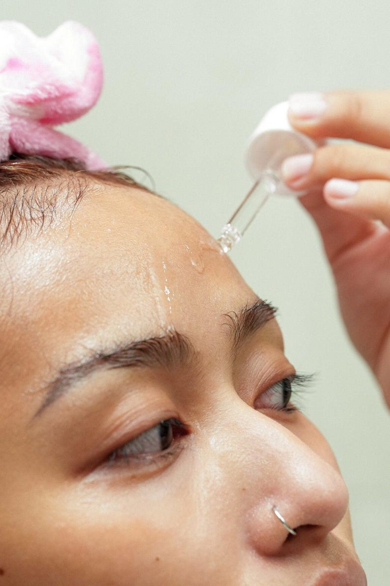 Close-up of model using dropper to apply Ruby Vibe Co Radiant Boost Serum on forehead, enhancing natural glow.